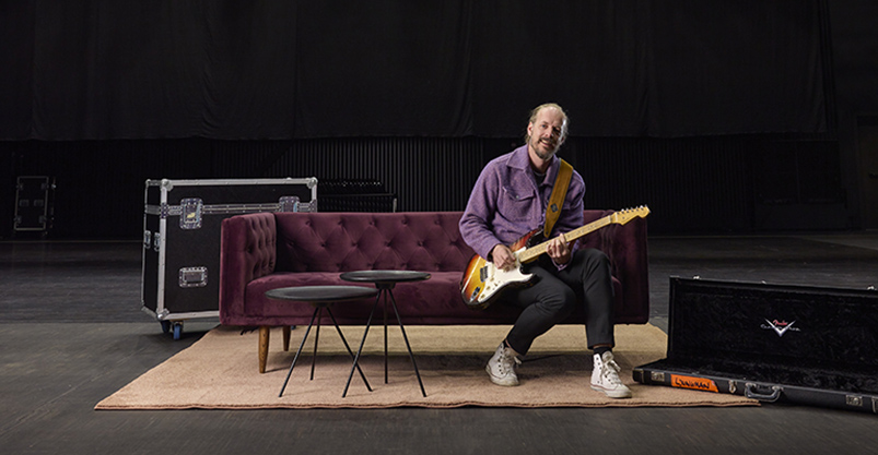 Man sitter på en stor svart låda. Han håller i en gitarr